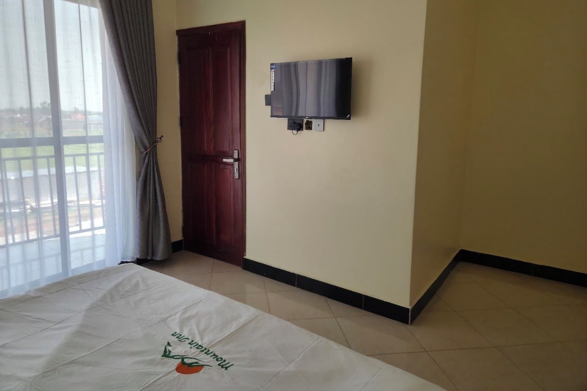 A room with a bed, a door, a wall-mounted TV, and a window with curtains.