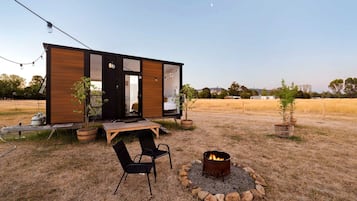 Uma cabana moderna com deck de madeira, duas cadeiras, lareira e um pequeno jardim.