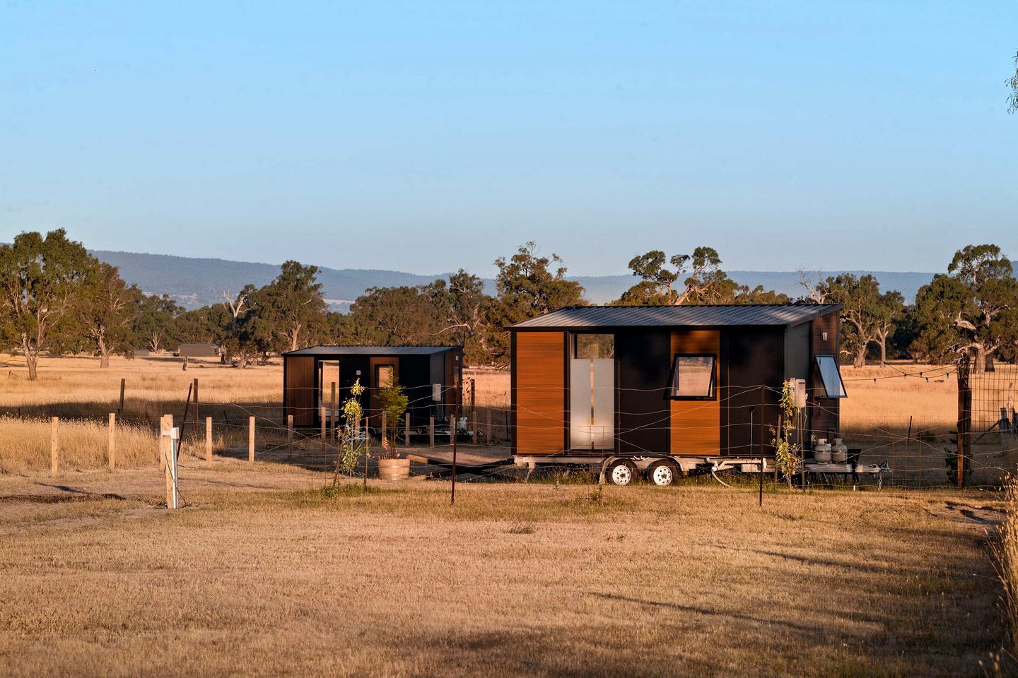 Two small, modern cabins with metal roofs, surrounded by a fenced area and dry grassland, with trees and a distant mountain range in the background.