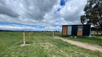 A small cabin with a window and a door, situated in a grassy field with a dirt path leading to it.