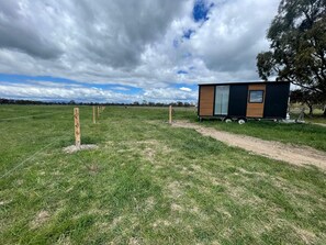 A small cabin with a window and a door, situated in a grassy field with a dirt path leading to it.