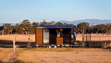 Een kleine, moderne blokhut met een metalen dak, gelegen in een landelijk veld met een heldere lucht en verafgelegen bergen.