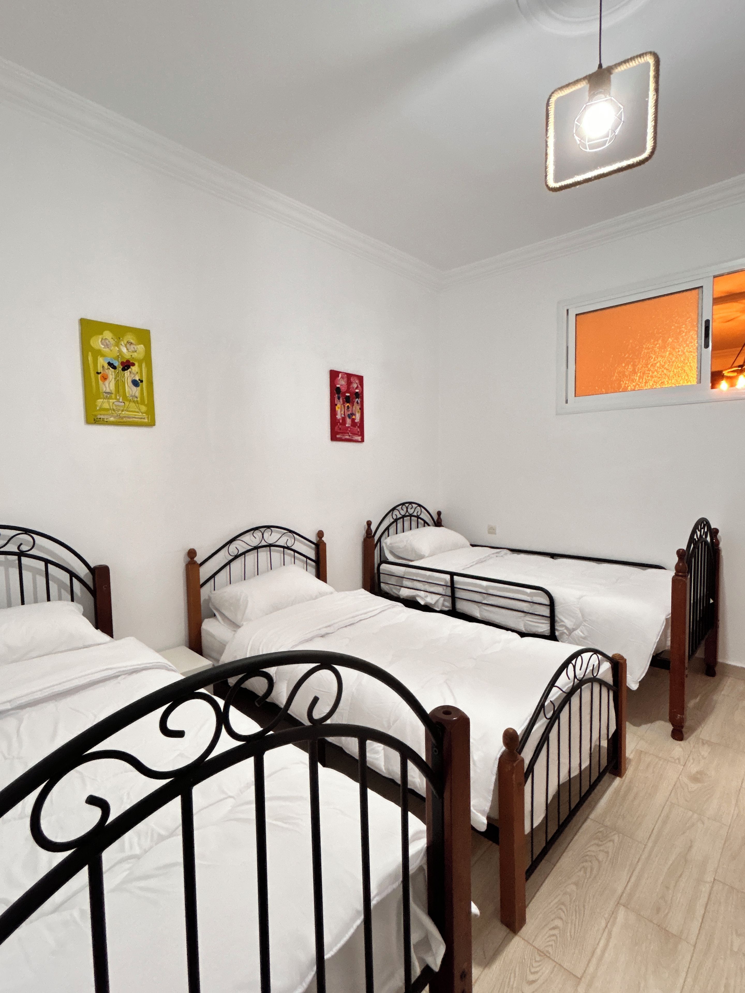 Une chambre avec trois lits simples, une petite fenêtre donnant sur un paysage agréable et un luminaire fixé au mur.