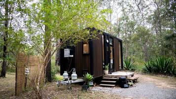 Une petite cabane en bois sombre, dotée d’un porche et entourée d’arbres et de végétation.