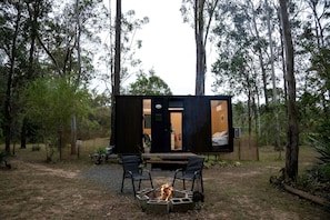 A small, modern cabin with a fire pit and two chairs in a forested area.
