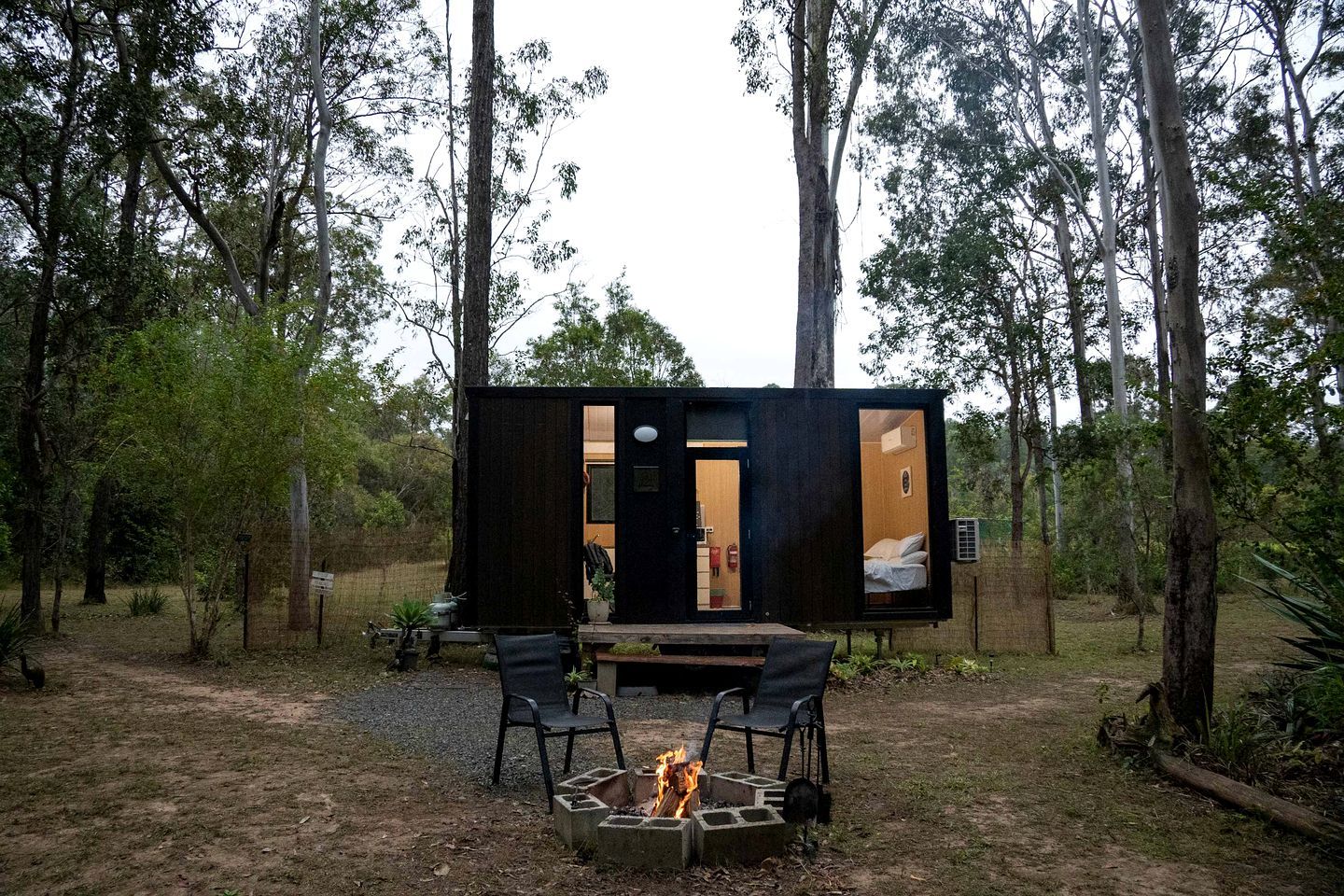 Una cabaña pequeña y moderna con un brasero y dos sillas en un área boscosa.