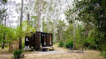 Une petite cabane de couleur sombre, située dans une zone boisée, avec un chemin de gravier menant à elle.