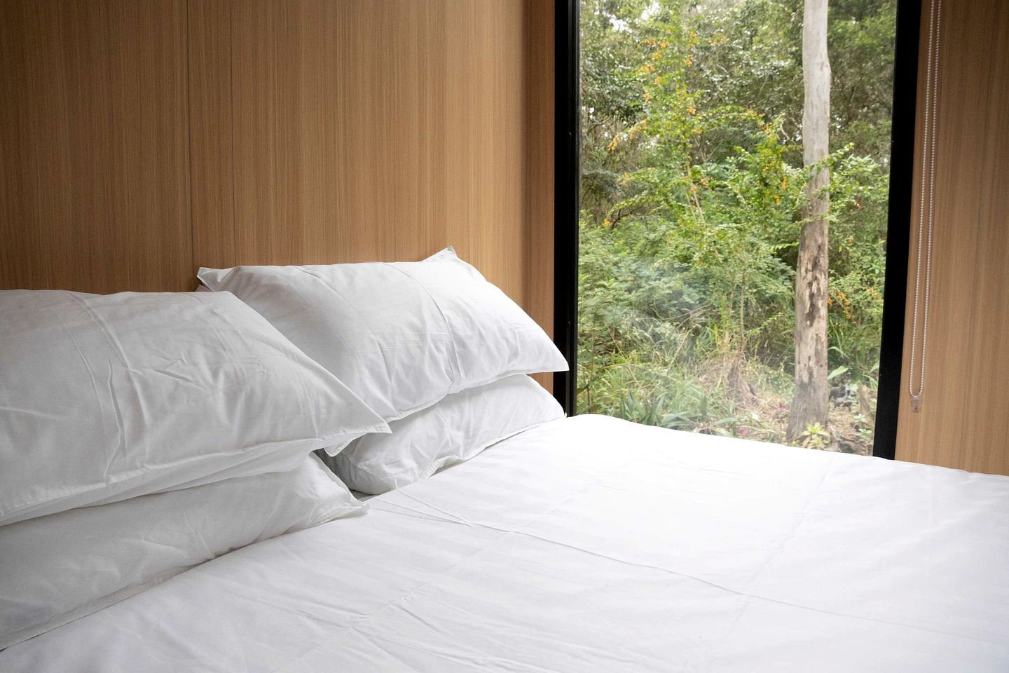Cama com lençóis brancos em quarto com painéis de madeira e janela com vista para área verde.