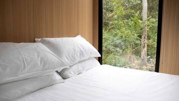 Cama com lençóis brancos em quarto com painéis de madeira e janela com vista para área verde.