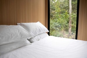 A bed with white linens in a room with wooden paneling and a window overlooking greenery.
