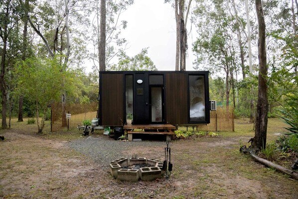 A small, dark wooden cabin with a bench outside, surrounded by trees.