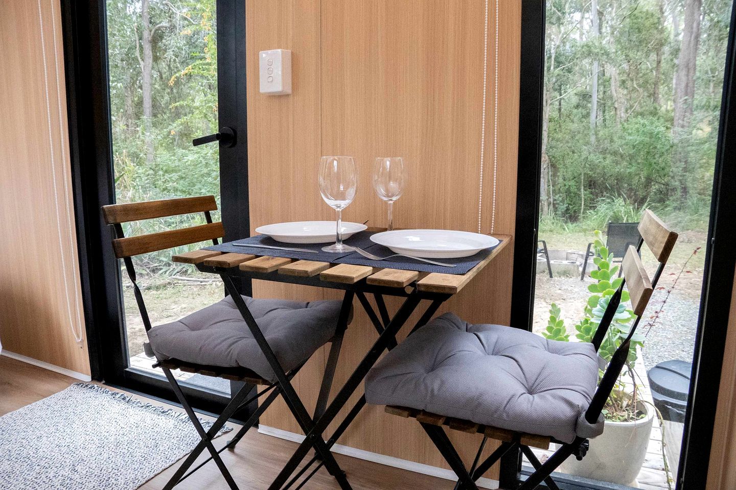 A dining area with a table set for two, two chairs, a bench, and a view of the outdoors.