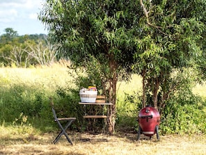 A picnic setup with a grill, a basket, and a folding chair in a grassy area.