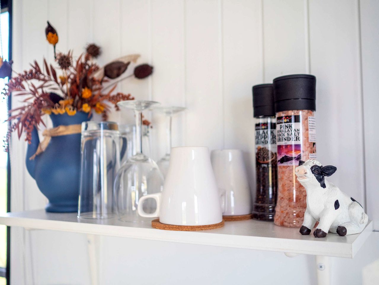 A shelf with white mugs, a glass pitcher, a blue vase with dried flowers, and salt grinders.