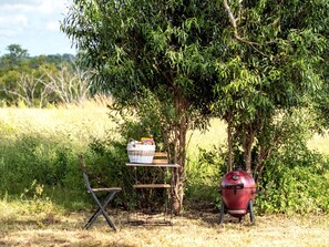 A picnic setup with a grill, a basket, and a folding chair in a grassy area.