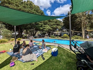 Un grupo de personas sentadas sobre una manta junto a una piscina, con una vela de sombra arriba.