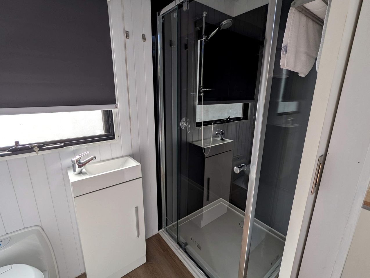 Ein modernes Badezimmer mit einer Duschkabine aus Glas, einem weißen Waschbecken und einem Fenster mit Jalousie.