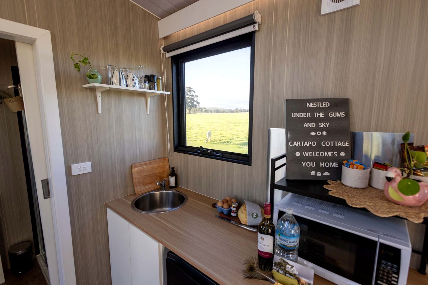 A compact kitchenette with a window, a sink, a microwave, and a sign welcoming guests to Cartapo Cottage.
