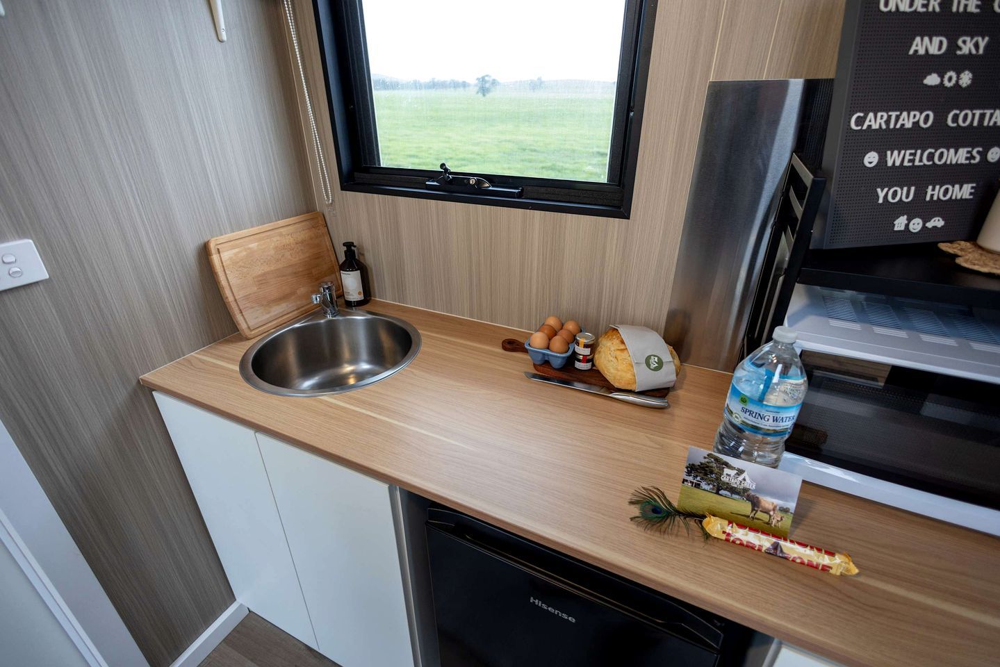 Een kleine keuken met een RVS spoelbak, een houten aanrechtblad en een raam met uitzicht op een veld.