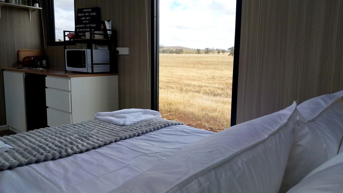 Ein Schlafzimmer mit einem Bett, einem Schreibtisch mit Computer und Blick auf ein Feld durch ein Fenster.