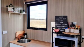 A kitchen with a window, a sink, a wooden chair, a shelf with kitchenware, and a sign welcoming guests.