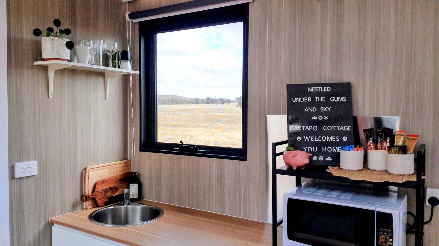 Une cuisine avec une fenêtre, un évier, une chaise en bois, une étag ère remplie d’ustensiles de cuisine et un panneau indiquant : « Bienvenue à nos invités ».