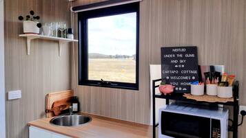 A kitchen with a window, a sink, a wooden chair, a shelf with kitchenware, and a sign welcoming guests.