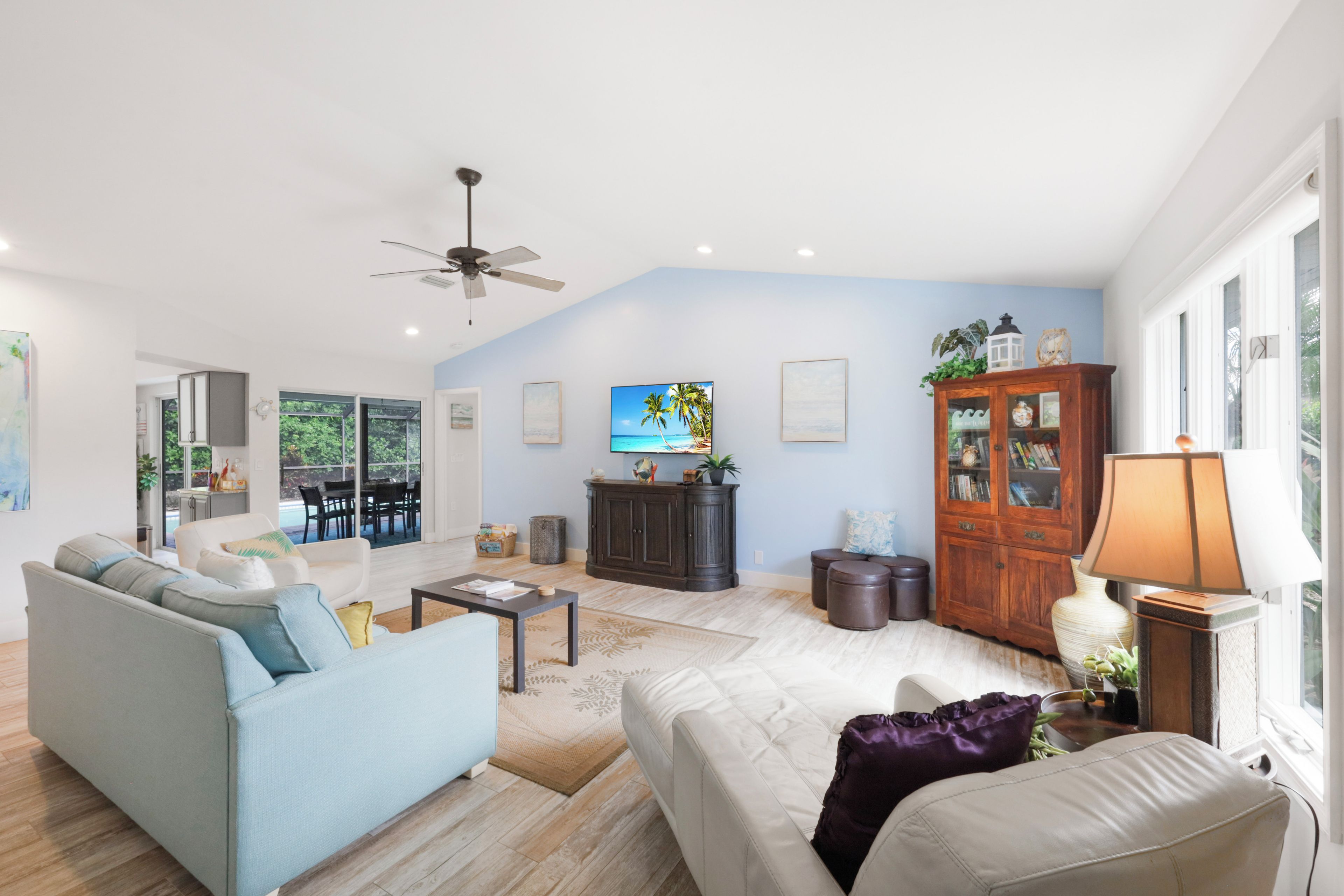 A spacious living room with a sofa, a coffee table, and a television. There is a ceiling fan, a cabinet, and a window with a view of the outdoors.