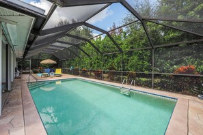 A covered swimming pool with a view of trees and a patio area with chairs and a table. - Private Inland Home | Heated Pool & Near Goodland (Marco Island)