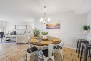 A dining area with a round table, white chairs, and a chandelier. - Fifth Floor Beachfront Condo | Pool, Gym & Pickleball (Marco Island)