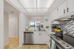 A modern kitchen with white cabinets, a stainless steel dishwasher, a black stove, and a tiled backsplash. - Fifth Floor Beachfront Condo | Pool, Gym & Pickleball (Marco Island)