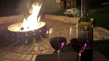 A table with two wine glasses and a bottle, a fire pit, and a pool in the background.
