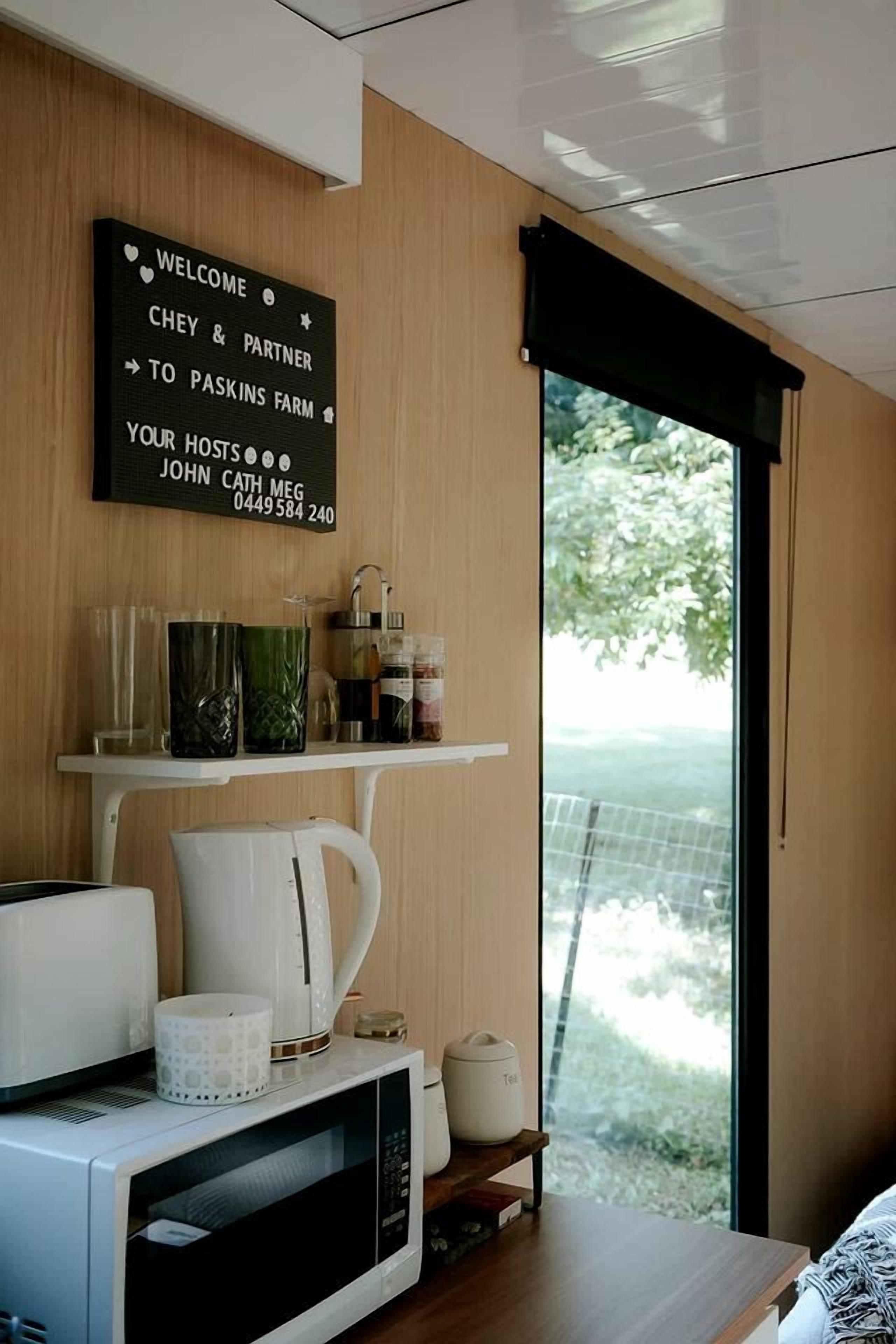 A modern room with a wooden wall, a welcome sign, a coffee machine, a kettle, and a view of greenery outside.