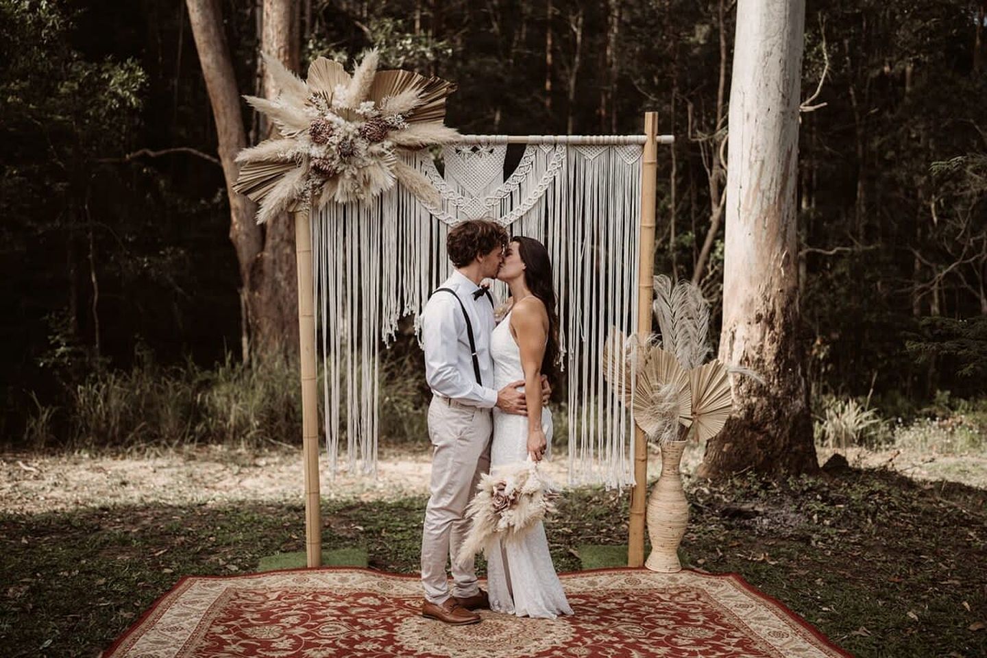 A couple standing in front of a macrame backdrop with a red patterned carpet.