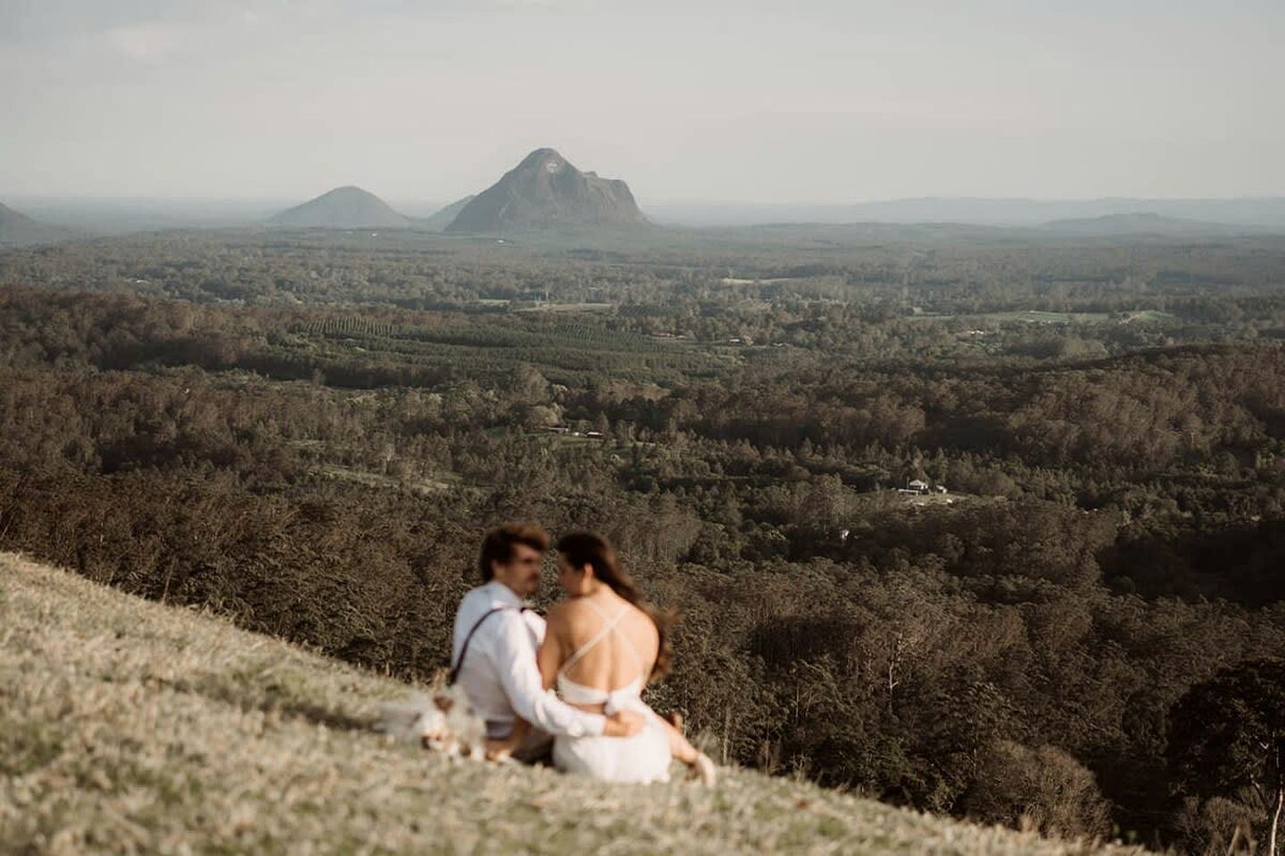 Un couple est assis sur une colline herbeuse, d’où l’on aperçoit une vallée boisée et, au loin, des montagnes.