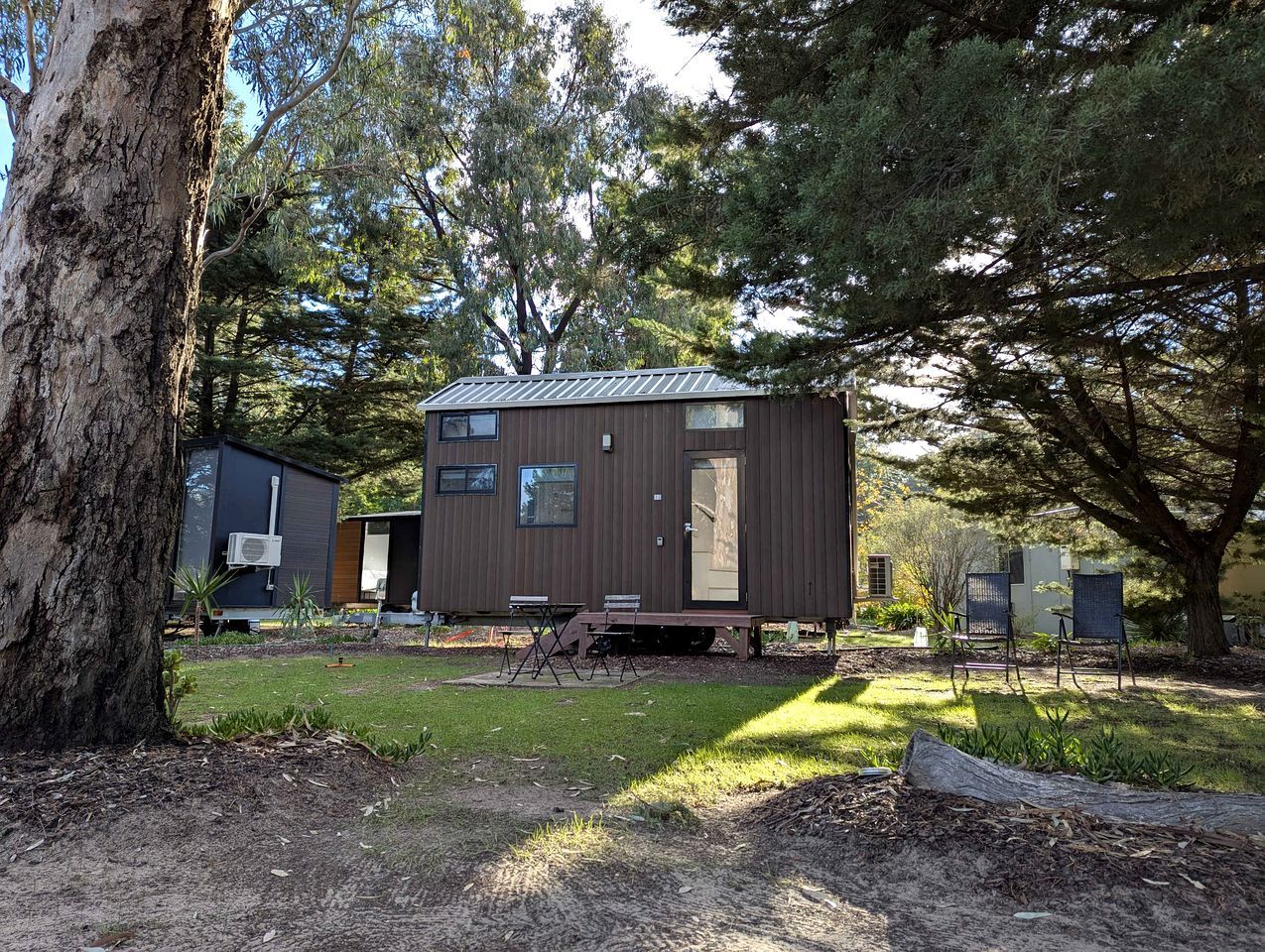 A small, single-story cabin with a metal roof, surrounded by trees and a grassy yard.