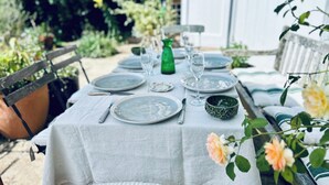 Mesa de jantar ao ar livre posta com pratos, talheres e copos, cercada por vegetação e um edifício branco ao fundo.