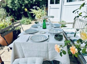 Et udendørs spisebord dækket med tallerkener, bestik og glas, omgivet af grøn vegetation og en hvid bygning i baggrunden.