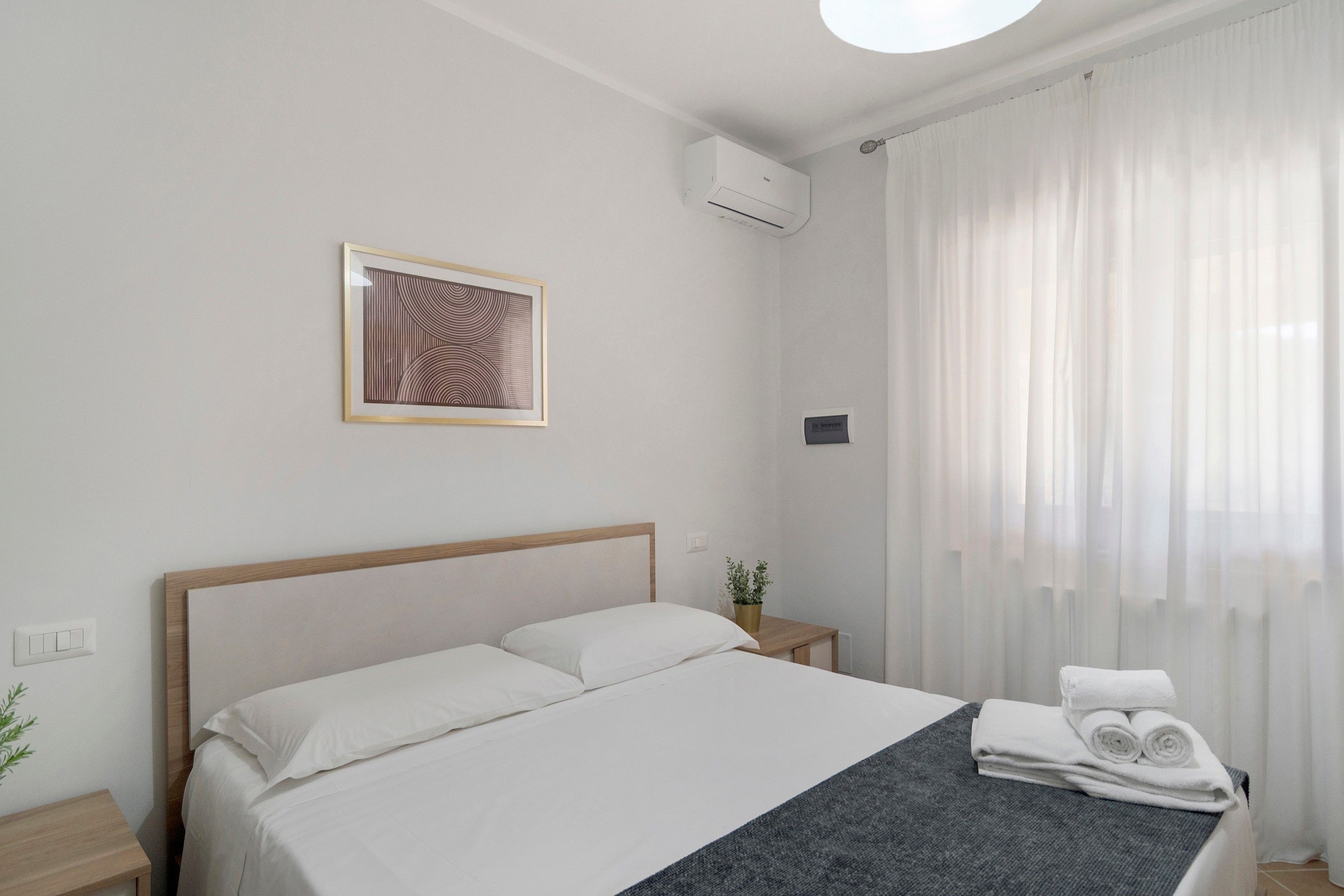Habitación de hotel con cama, un cuadro enmarco, un equipo de aire acondicionado, una planta pequeña y una ventana con cortinas.