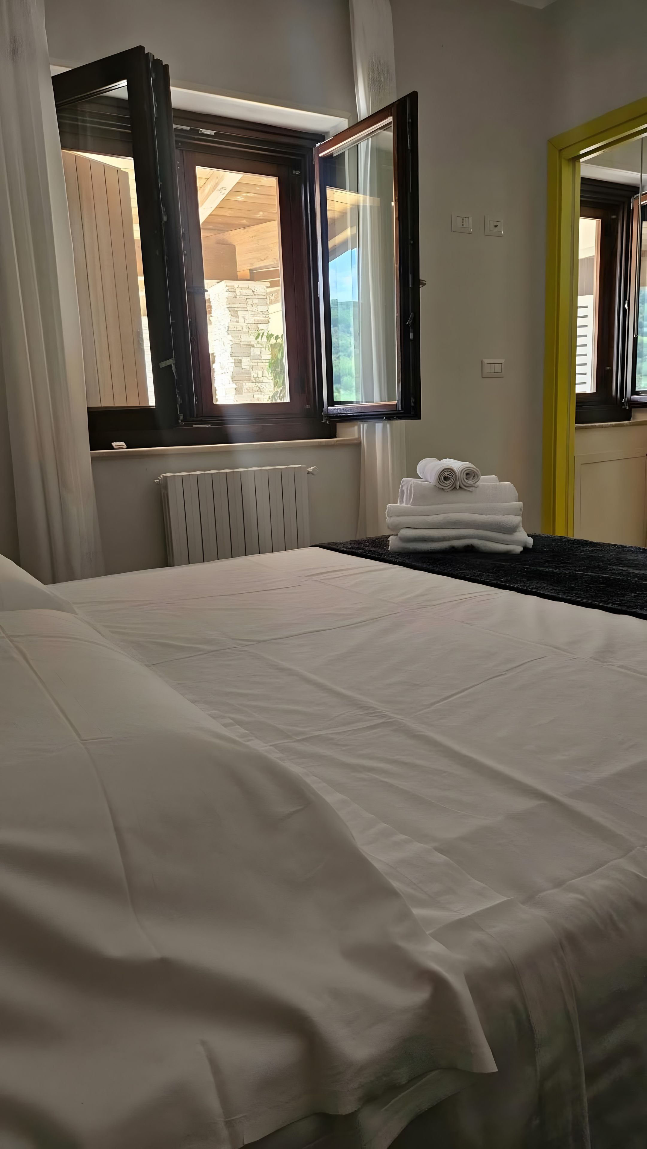 Una camera d'albergo con un letto, un termosifone, una finestra con tende e un mucchio di asciugamani.
