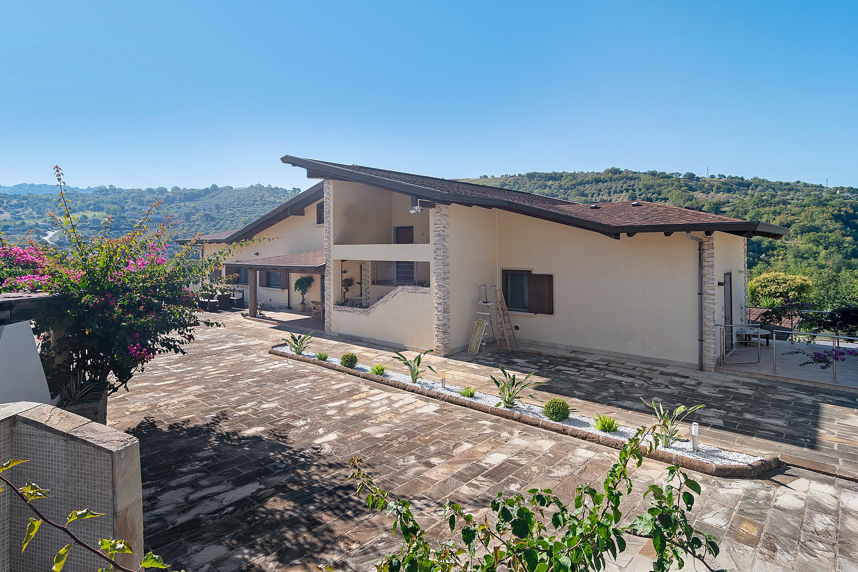A two-story building with a flat roof, stone pillars, and a paved courtyard with plants.