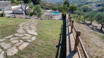 A stone pathway leading to a pool area with a wooden fence, surrounded by greenery and hills.