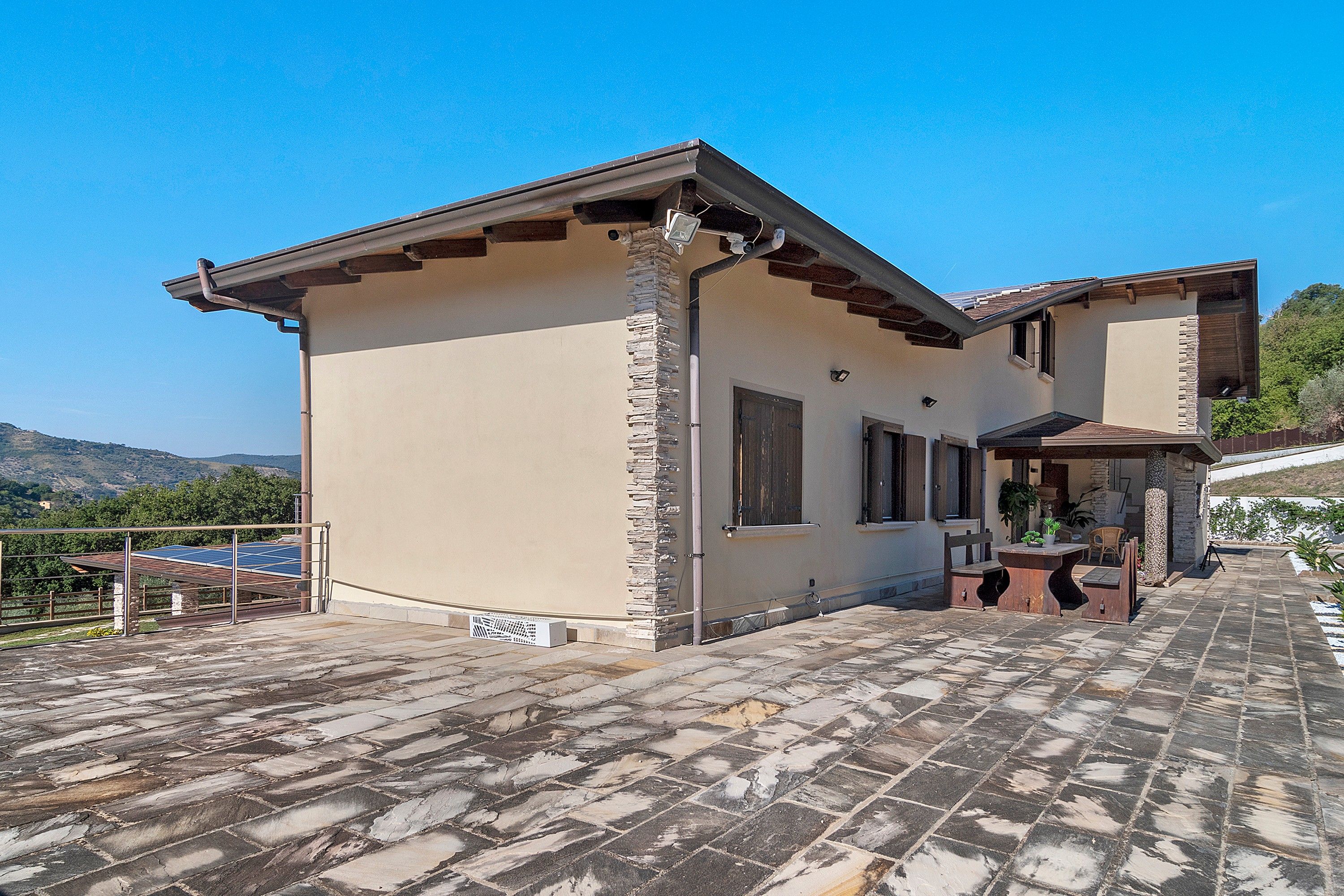 Una casa de dos pisos con fachada de piedra, techo plano y un patio con mobiliario exterior.