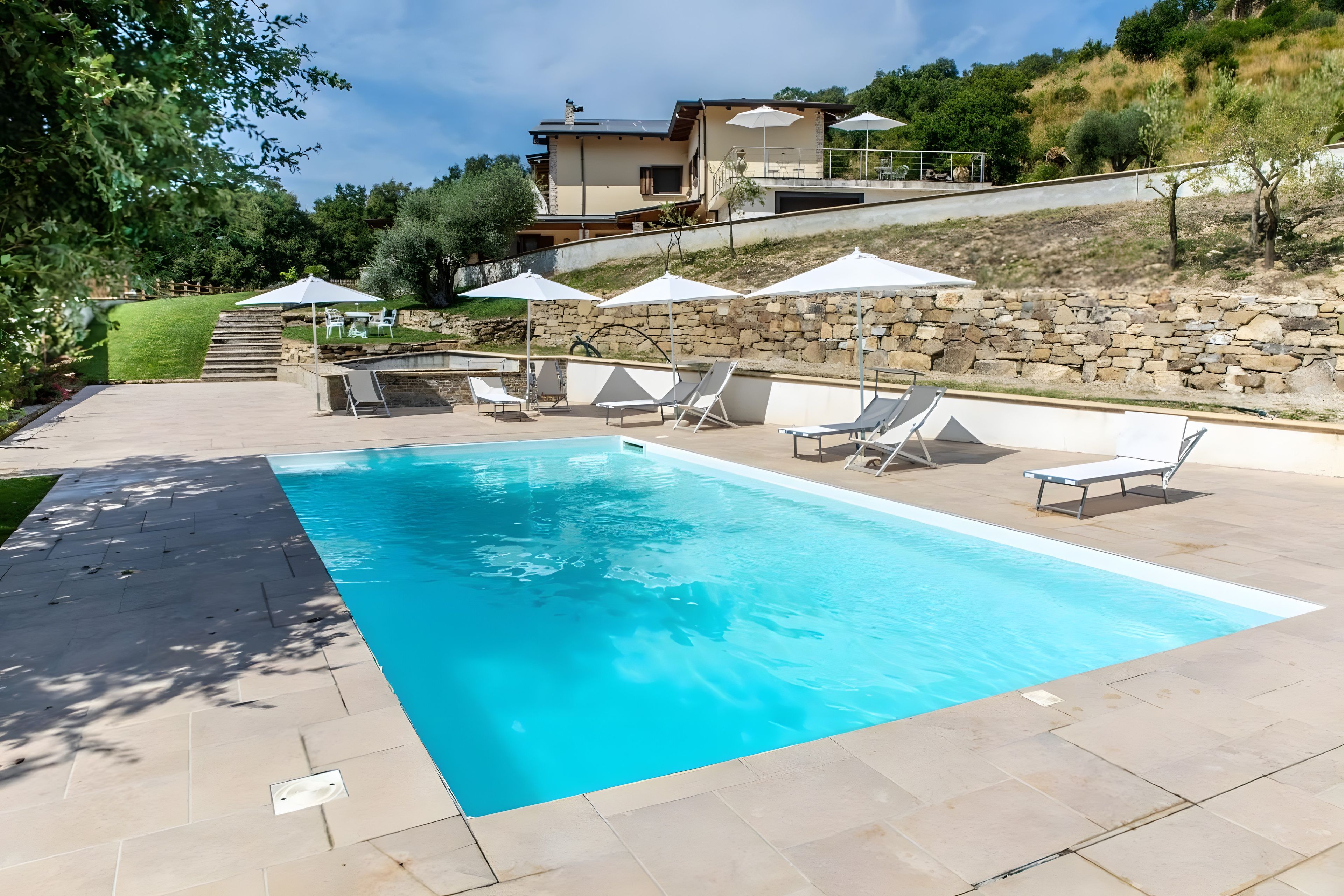 Una piscina rectangular con un muro de piedra y una casa al fondo.