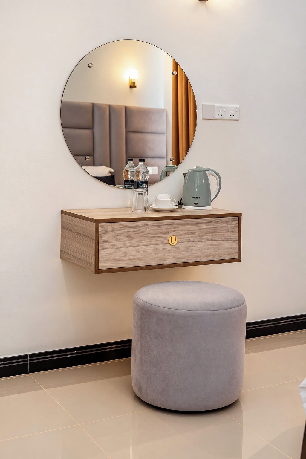 A modern bathroom vanity with a round mirror, a wooden cabinet, a grey ottoman, and a coffee maker.