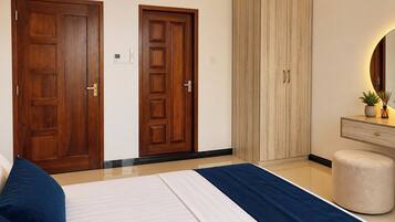 Un dormitorio con una cama, dos puertas de madera, un armario, una tocador con espejo y un pequeño otoman redondo.