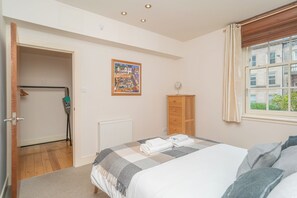 Een slaapkamer met een bed, een radiator, een kledingkast, een deur naar een andere kamer en een raam met uitzicht op gebouwen.