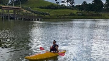 A person kayaking on a calm lake with a grassy hill and a wooden dock in the background.