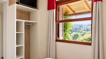 A bedroom with a bed, a TV mounted on the wall, a window with curtains, and a view of greenery outside.