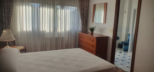 Un dormitorio con cama, cómoda, lámpara y ventana con cortinas.
